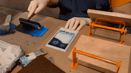 A video showing the process of applying ink to a printing plate with a roller, putting a card on the plate, pressing down on the paper, and eventually removing it showing the image on the paper.